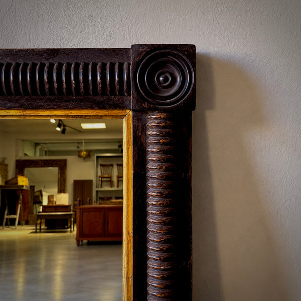 Close-up of an ornate dark brown carved wooden mirror frame with a spiral column detail and a gold-toned inner border, set against a light gray wall with a showroom interior visible in the mirror reflection.