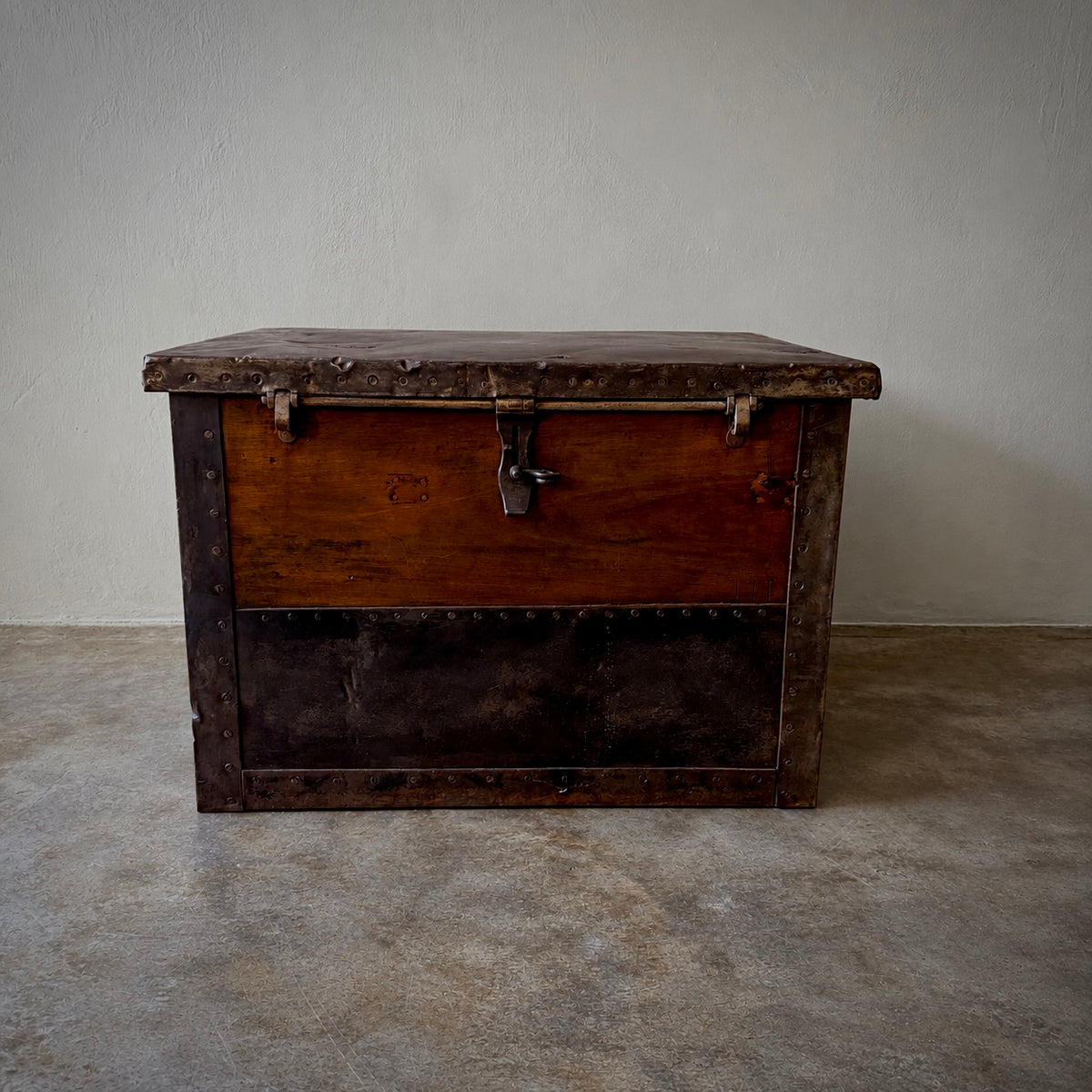 A vintage wooden storage chest with dark metal trim and a latch, photographed against a plain light wall and concrete floor.