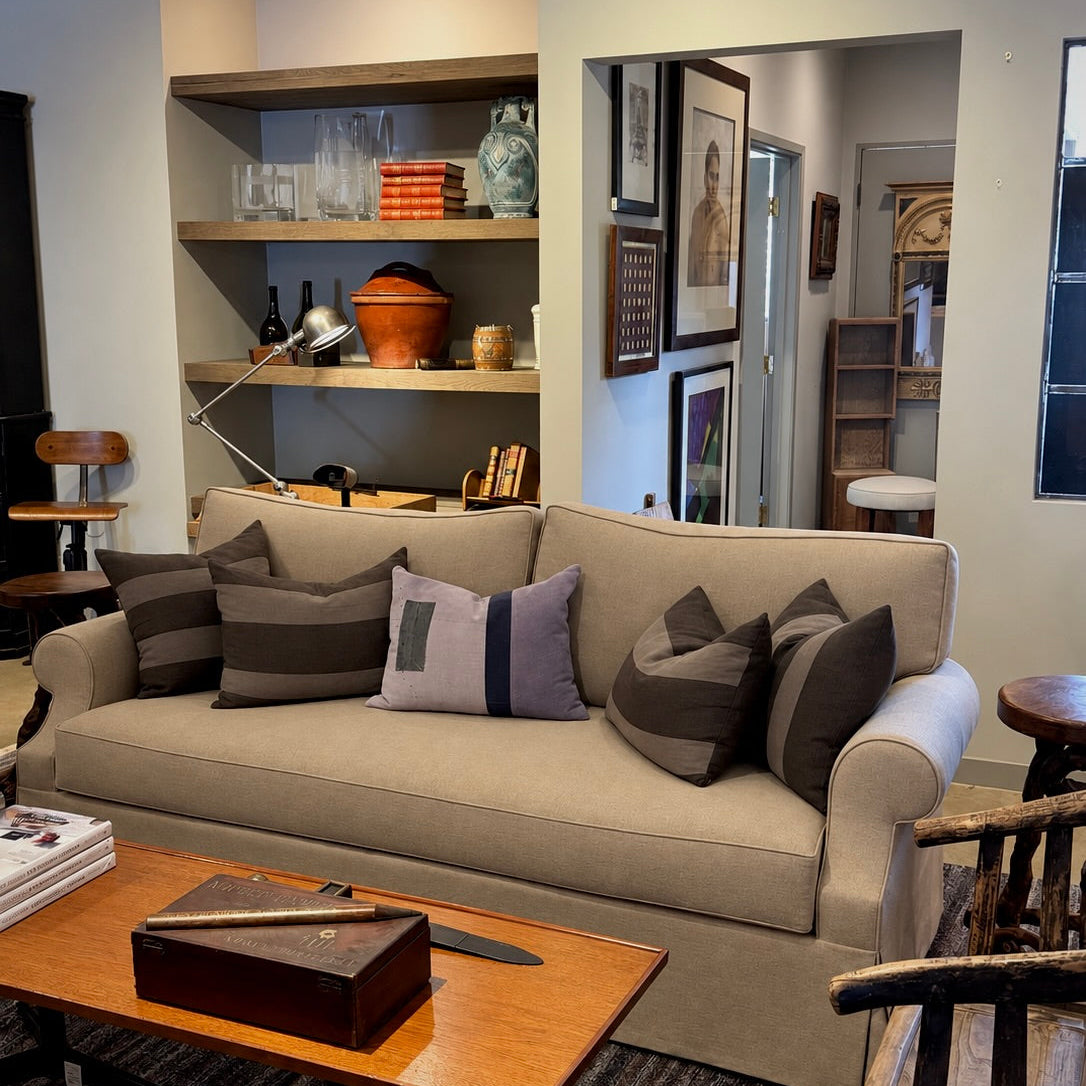 A beige upholstered sofa in a styled living room setting, decorated with multiple dark gray and striped throw pillows, positioned next to a wooden coffee table and shelves in the background.