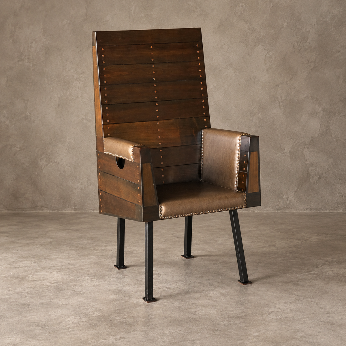 A rustic industrial-style accent chair with a tall dark brown wooden slat backrest, brown leather seat and armrests, brass nailhead trim, and black metal legs, set against a textured gray concrete wall and floor.