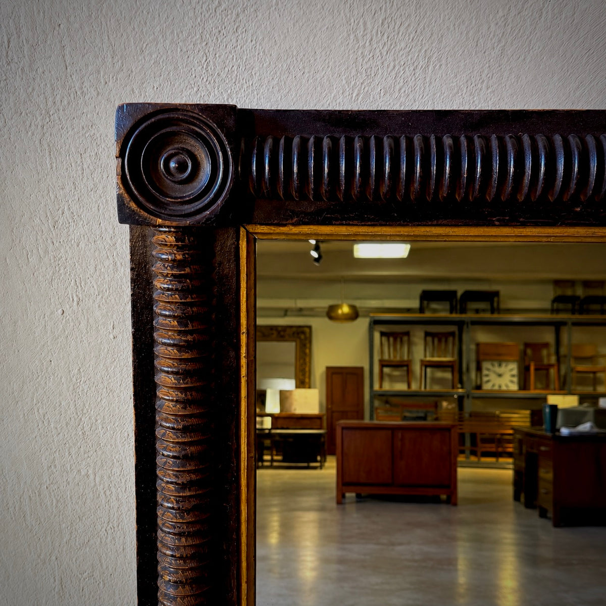 Close-up of an ornate dark wood mirror frame with carved details, leaning against a textured beige wall and reflecting a furniture showroom interior.