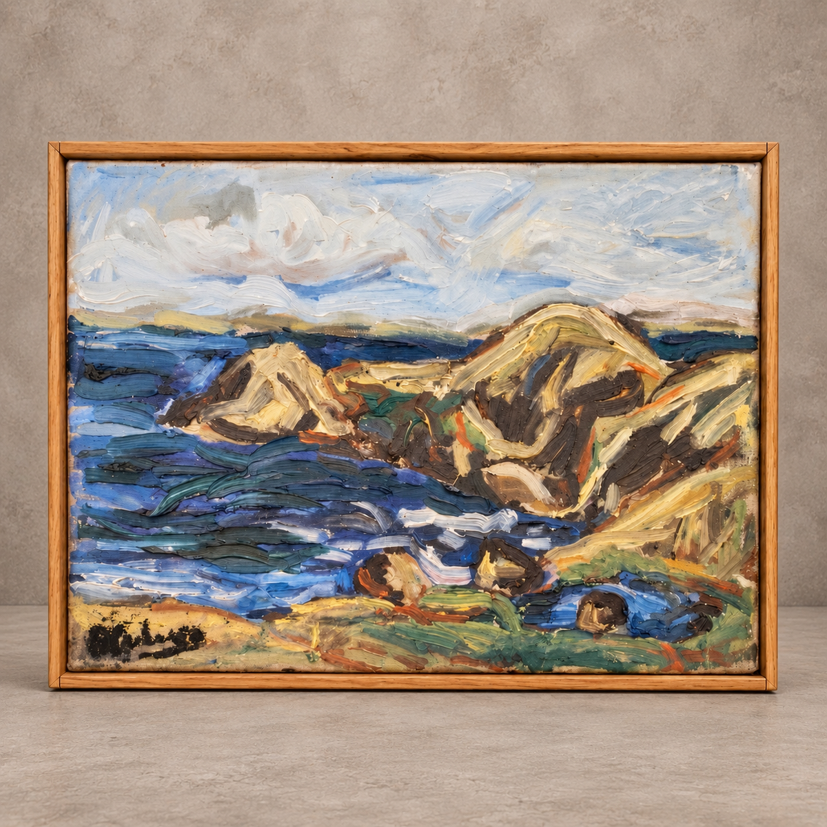 A framed landscape painting depicting a rocky coastline with blue ocean waves, tan and brown cliffs, and a cloudy sky, set in a light wood frame against a textured gray wall.
