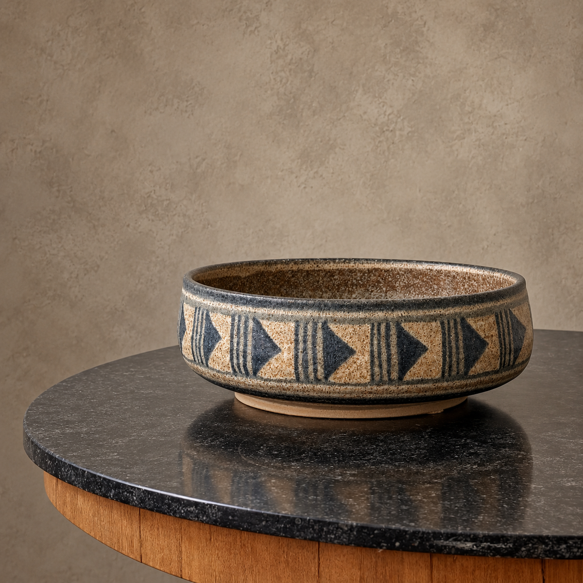 A round ceramic bowl with a Southwestern geometric pattern in dark blue and beige, displayed on a dark stone tabletop against a textured beige wall.
