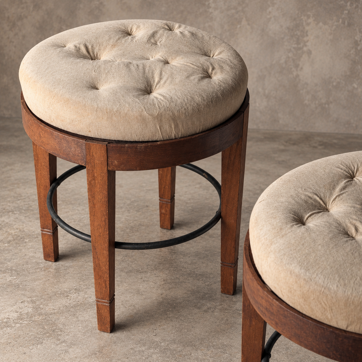 A pair of rustic circular wooden stool bases with four tapered legs and a black metal circular support ring and a rn upholstered top, photographed against a textured gray concrete backdeop