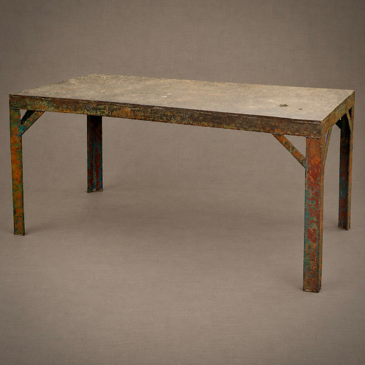 A rustic industrial metal table with a weathered, distressed finish showing rust, orange, and green patina over a gray base, set against a solid taupe background.