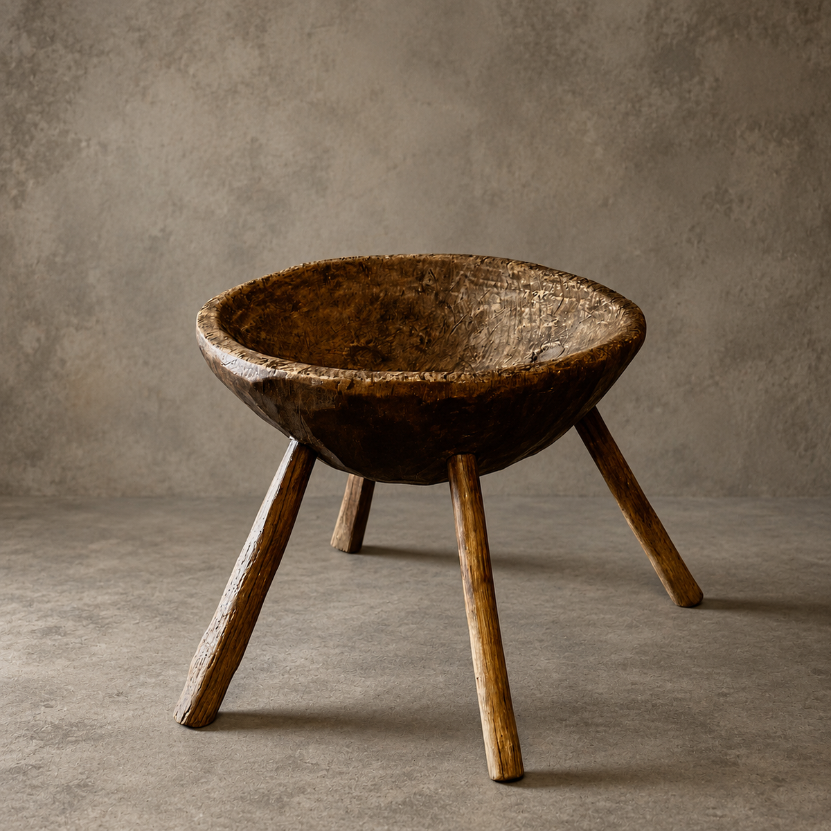 A rustic wooden bowl-shaped stool with three sturdy legs, photographed in a studio setting against a textured gray concrete wall and floor.