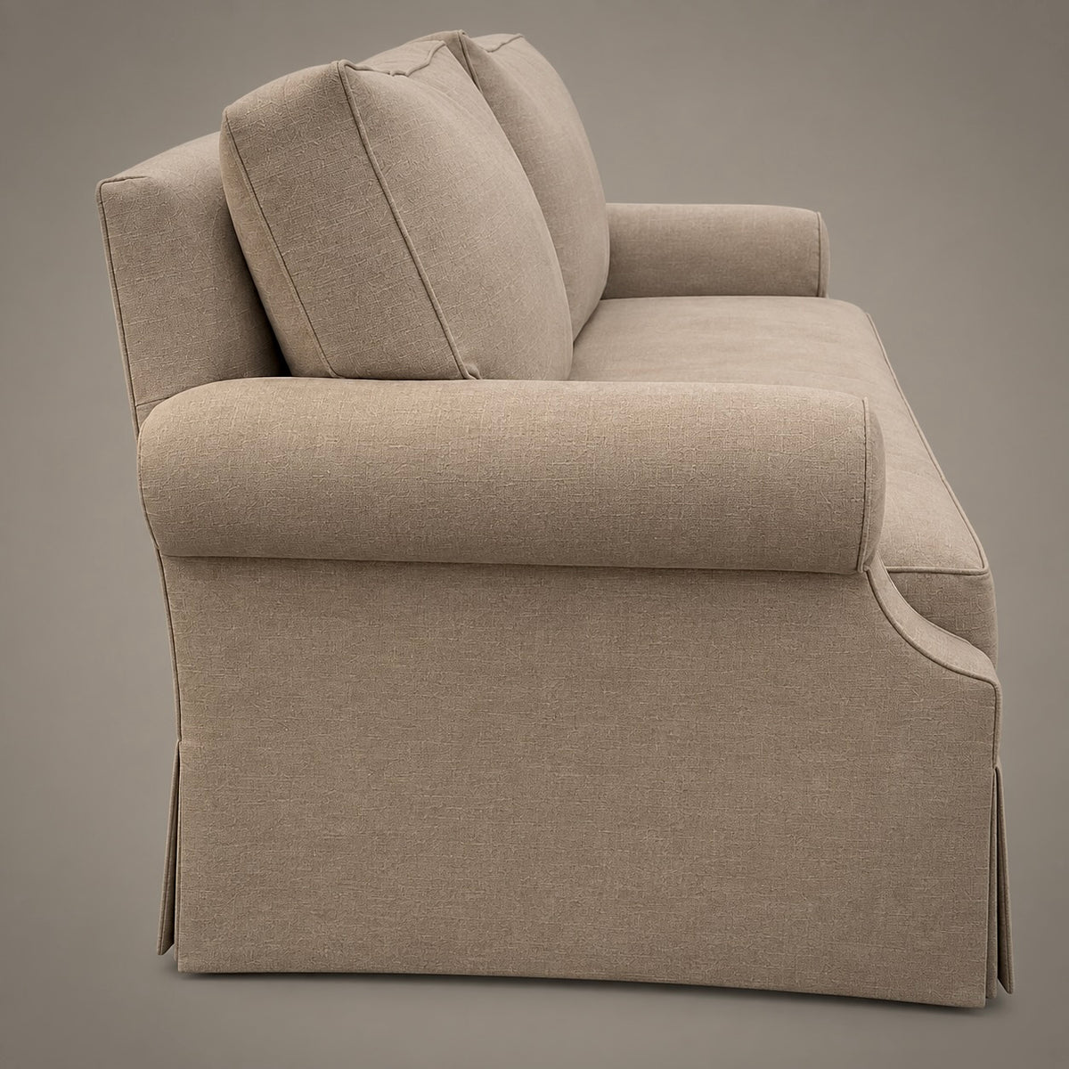 A beige upholstered sofa shown from a three-quarter side angle against a solid taupe-gray studio background.