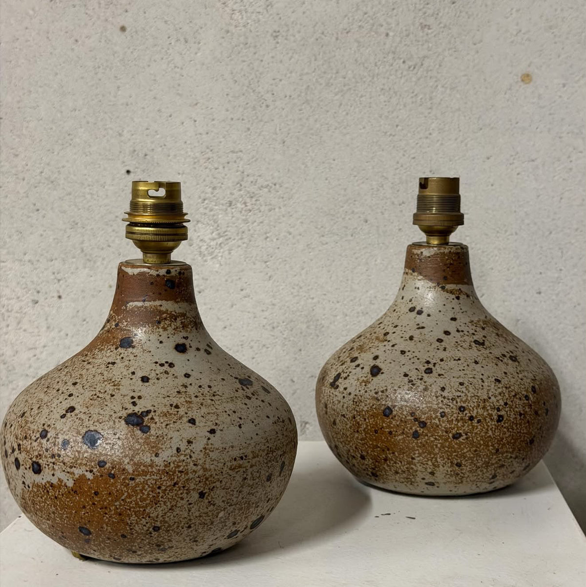 Two handmade ceramic table lamps with rounded bulb-shaped bodies, speckled brown and beige glaze, and brass lamp sockets, displayed on a white shelf against a textured gray wall.