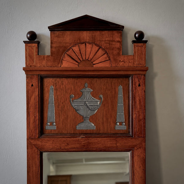 A vintage-style wooden wall mirror frame with a warm reddish-brown finish, featuring decorative inlays of a fanburst, an urn, and two obelisks, with a mirror reflecting a room interior.