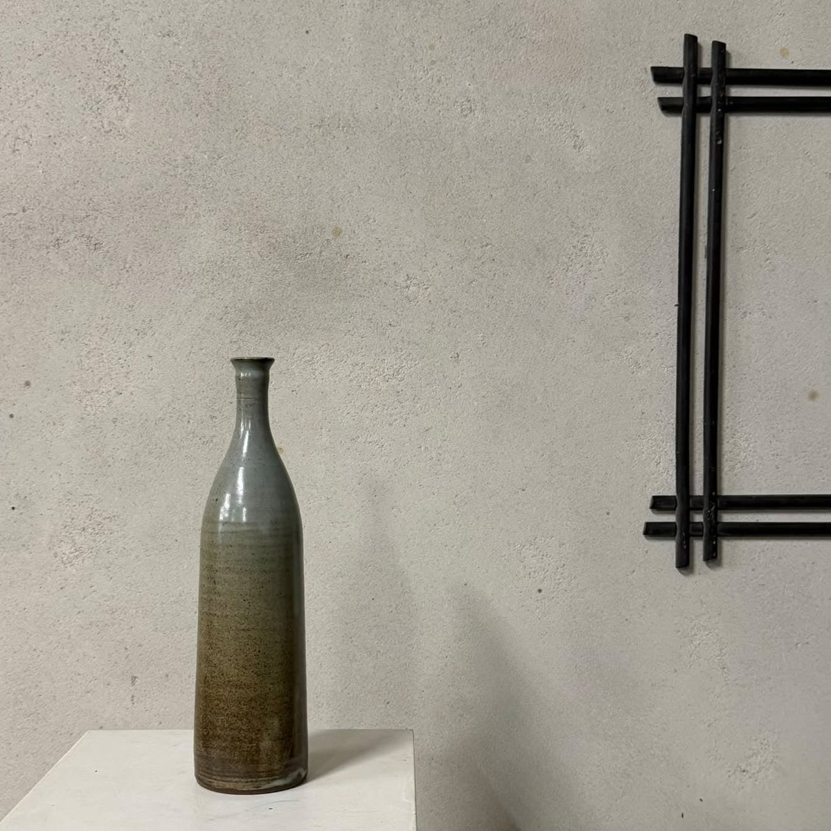 A tall, narrow ceramic vase with a textured glaze transitioning from gray-green at the top to earthy brown at the base, displayed on a white pedestal against a textured concrete wall.