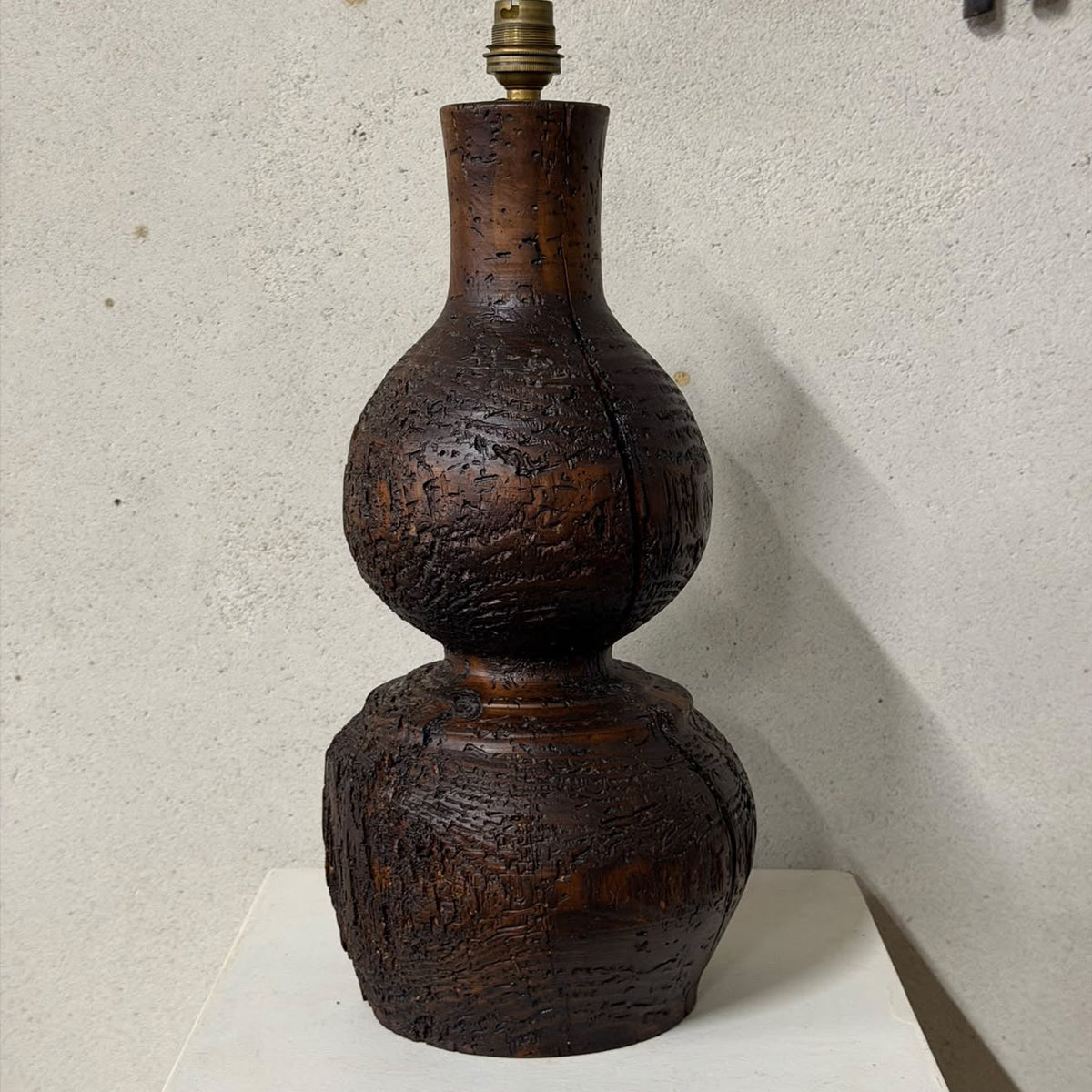 A dark brown carved wooden gourd lamp base with a textured, hand-hewn finish and a brass socket at the top, displayed on a white pedestal against a textured light gray wall.