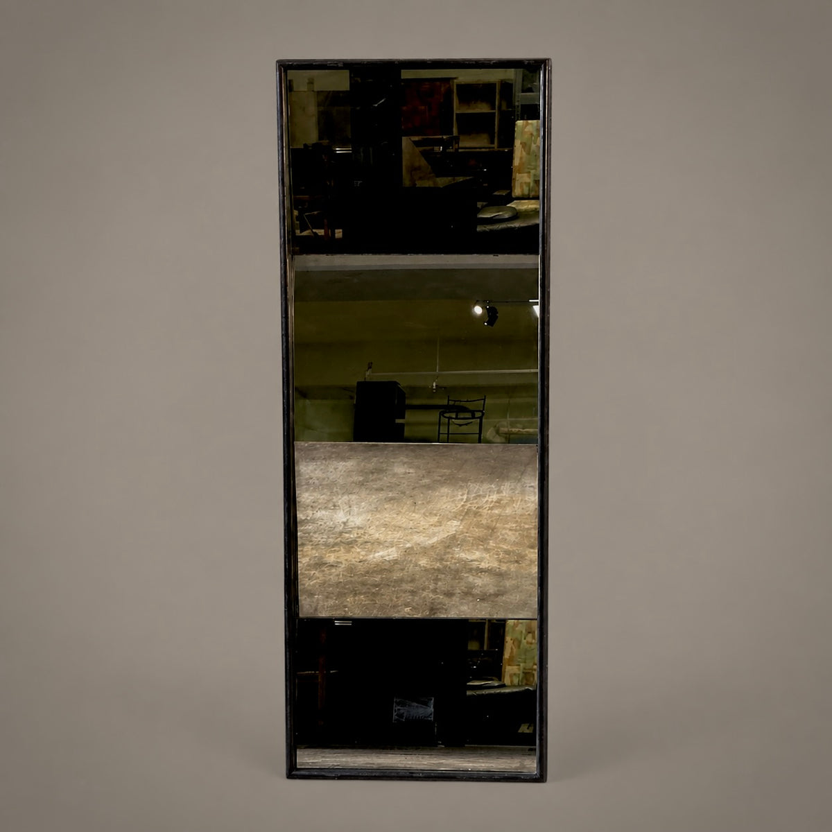 A tall, narrow rectangular mirror with a thin dark frame, reflecting an interior space with a prominent textured metallic panel in the center, set against a solid gray background.