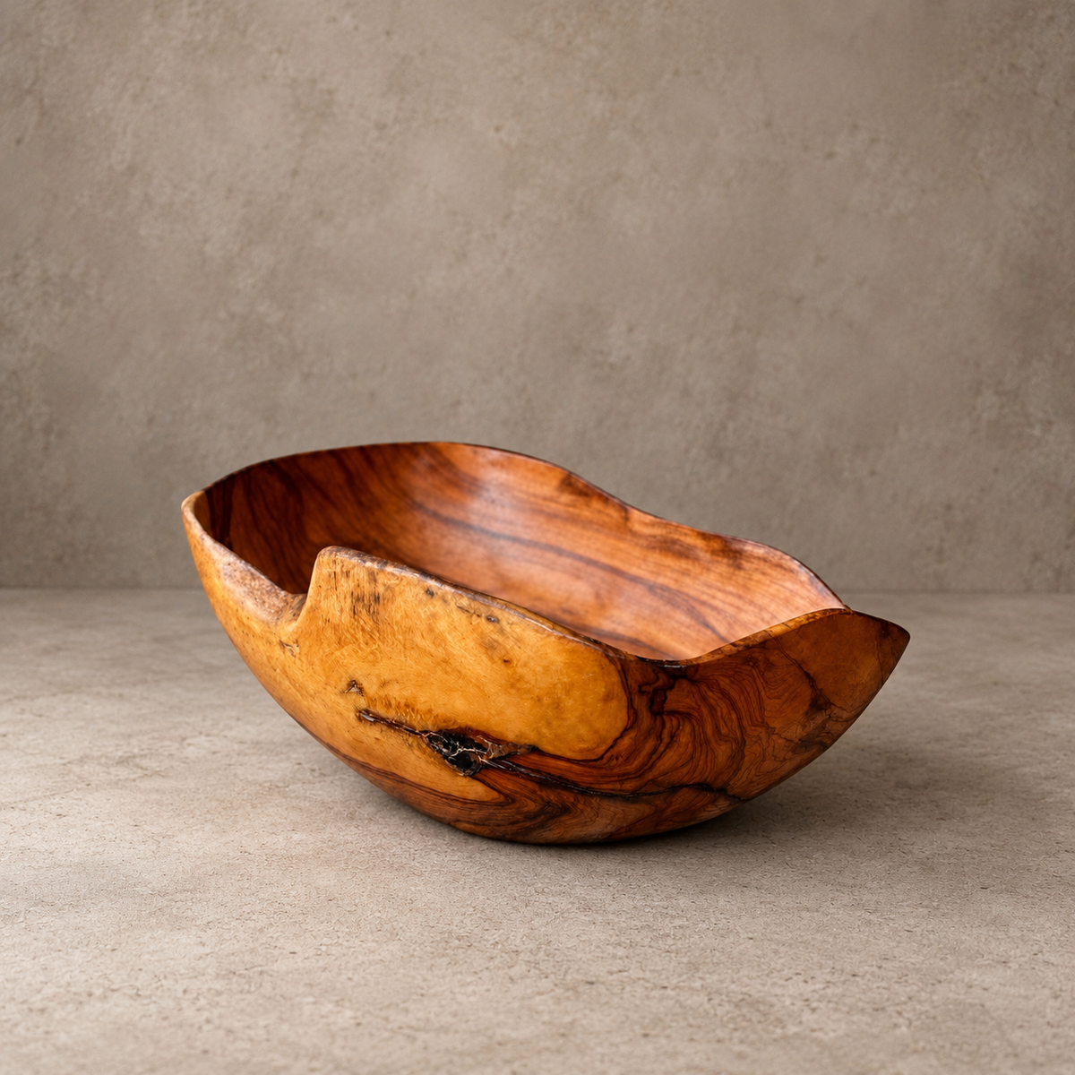 A rustic handcrafted wooden bowl with a wavy, organic shape and rich natural grain patterns, displayed on a neutral concrete surface against a textured gray wall.