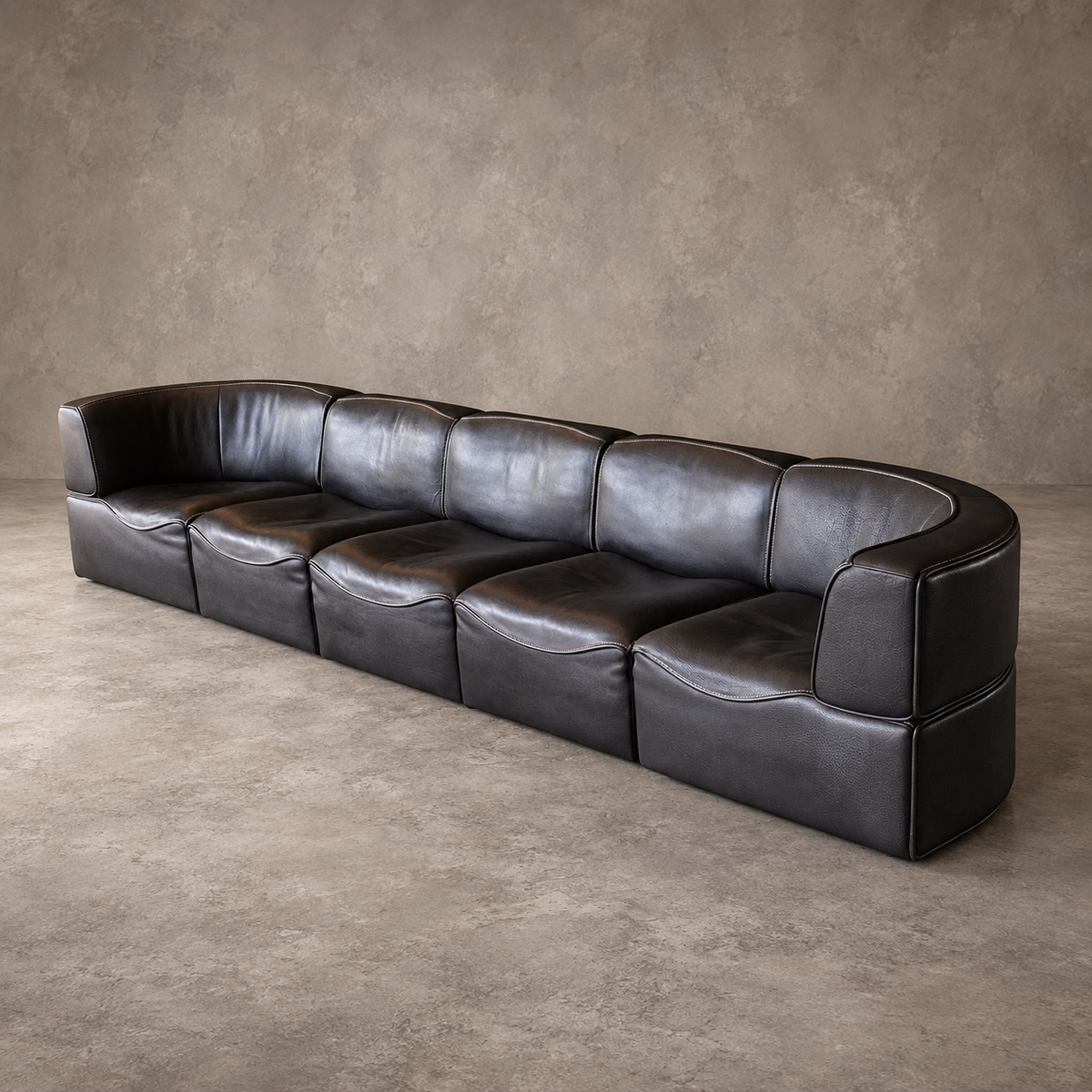 A long, curved modular sofa upholstered in dark charcoal gray leather, positioned on a textured concrete floor against a matching concrete wall.