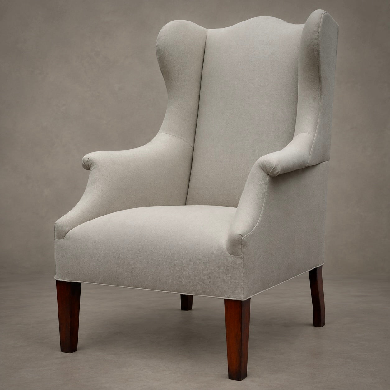 An upholstered wingback armchair with a light beige fabric and dark brown wooden legs, photographed against a neutral textured gray studio background.