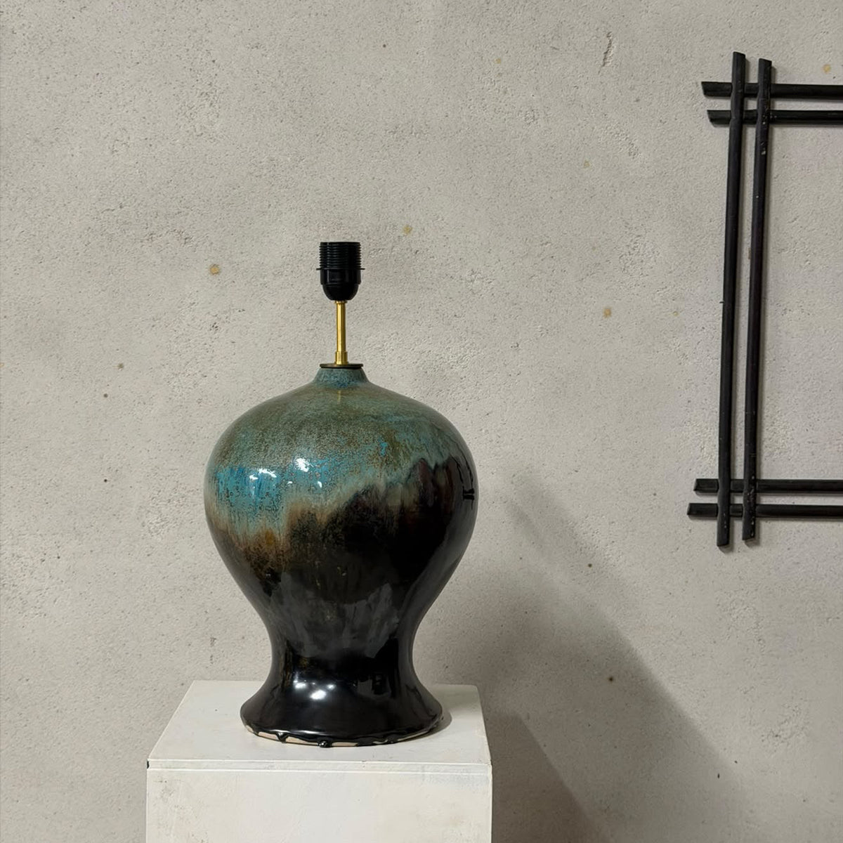 A ceramic table lamp with a rounded bulb-shaped body, featuring a glossy glaze that transitions from teal-blue at the top to deep black at the base, displayed on a white pedestal against a textured concrete wall.