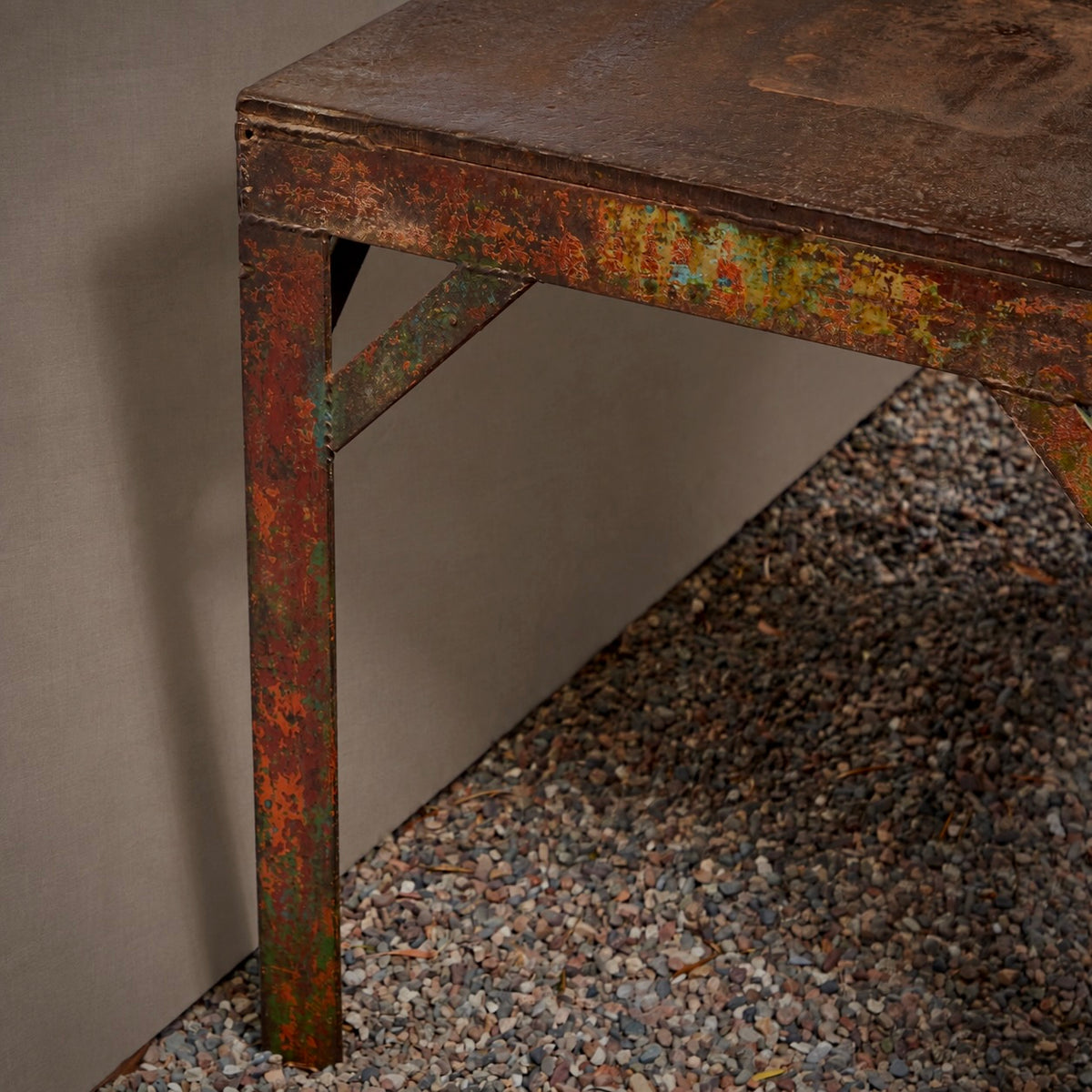 Close-up of a rustic metal table leg with a heavily distressed, rusted finish showing orange, green, and brown layers, positioned next to a plain wall and gravel ground.