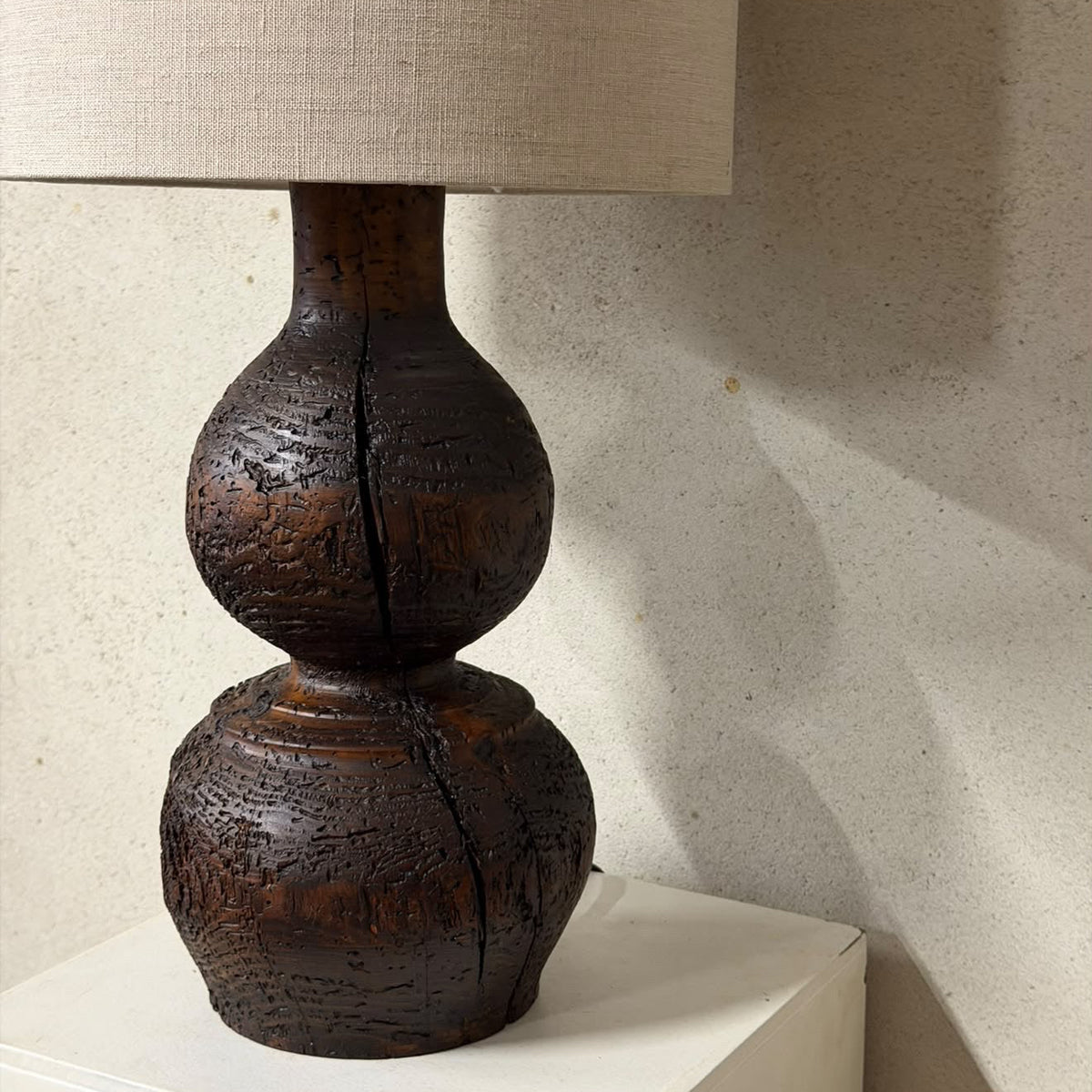 A rustic dark brown wooden table lamp with a textured, crackled finish and a beige linen drum shade, resting on a white pedestal against a textured beige wall.