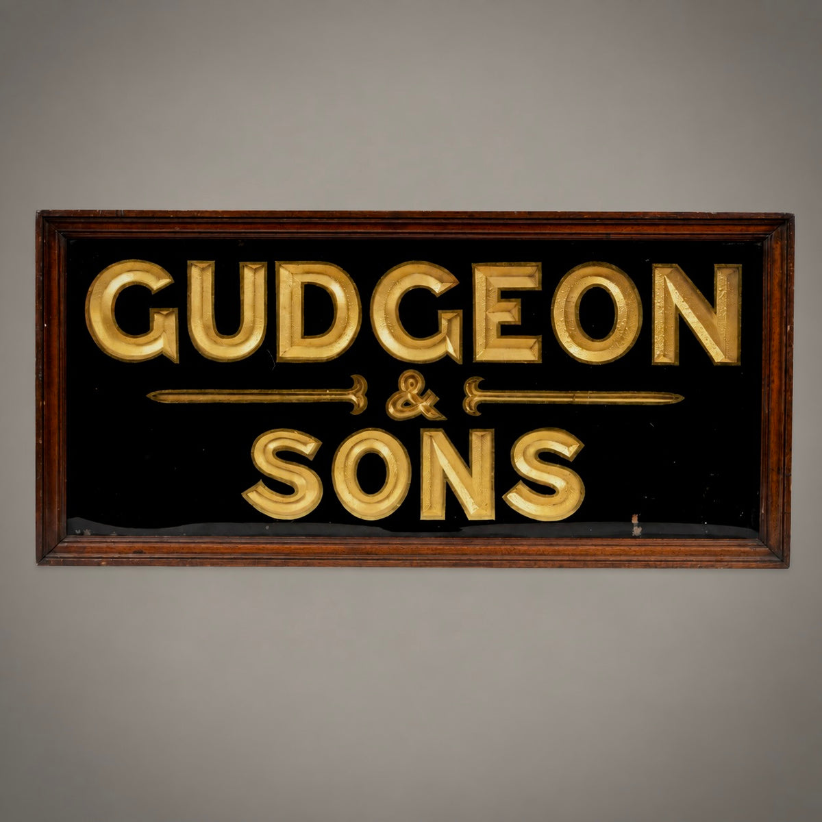 A vintage-style rectangular sign reading 'GUDGEON & SONS' in raised gold lettering on a black background, set in a dark wood frame against a plain gray wall.