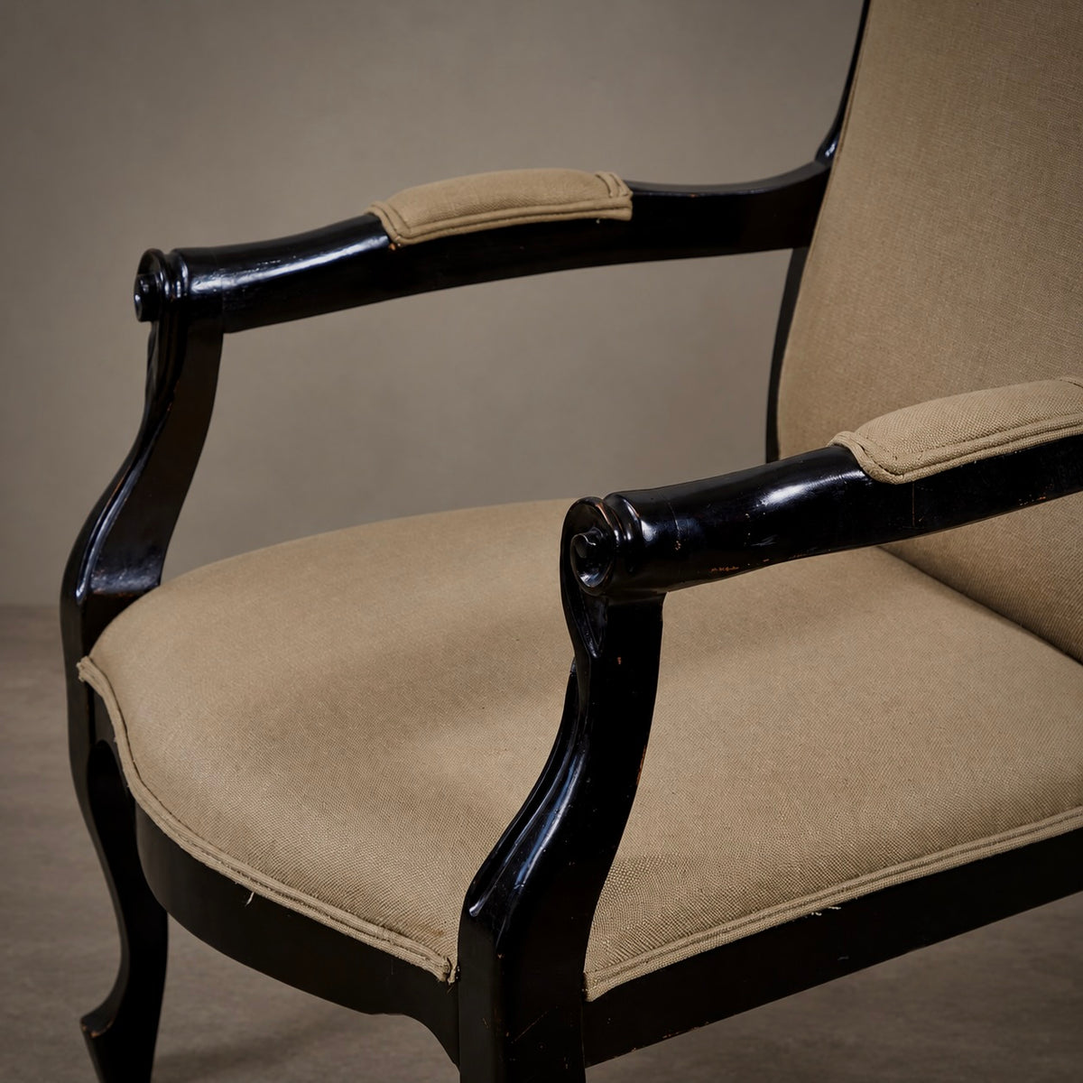 A close-up of a classic armchair featuring a glossy black carved wooden frame and beige fabric upholstery on the seat, backrest, and arm pads.