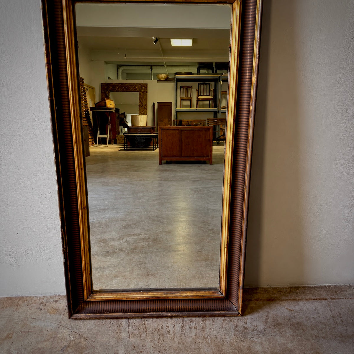 A tall rectangular floor mirror with an ornate dark brown and gold frame, leaning against a beige wall in an antique store or furniture showroom.