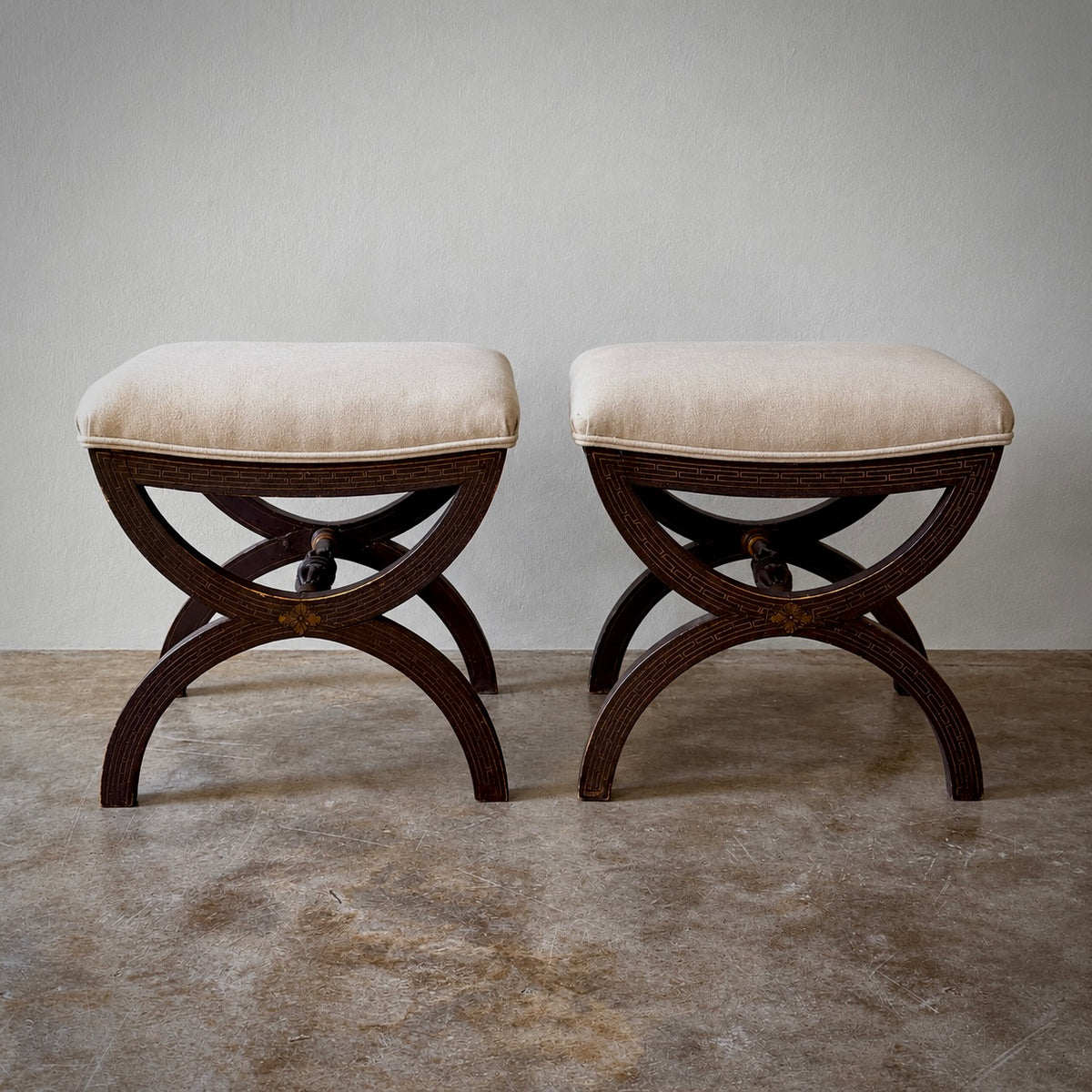 Two matching upholstered stools with cream fabric seats and dark carved wooden legs, shown side by side against a plain light gray wall and textured floor.