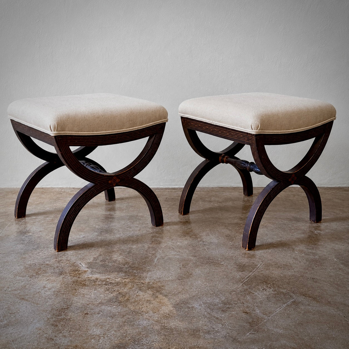Two matching upholstered ottomans with cream fabric seats and dark brown wooden legs, shown on a concrete floor against a light gray wall.