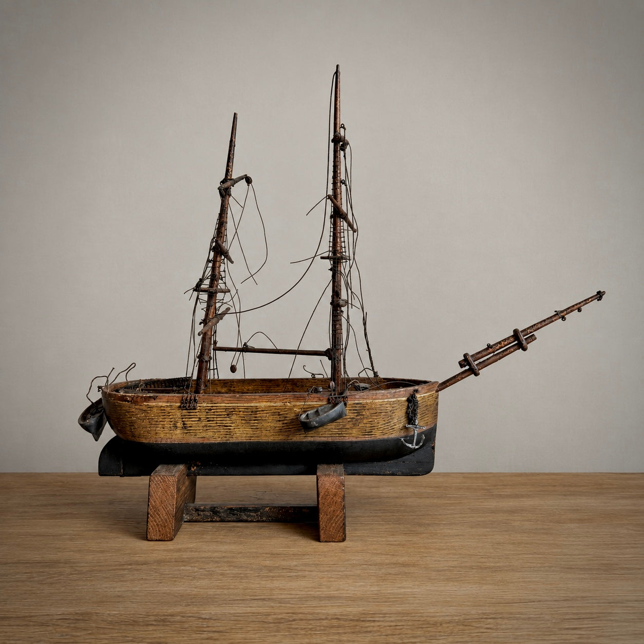 A vintage wooden model of a two-masted sailing ship with intricate rigging, mounted on a black base and supported by two wooden blocks, set against a neutral beige background.