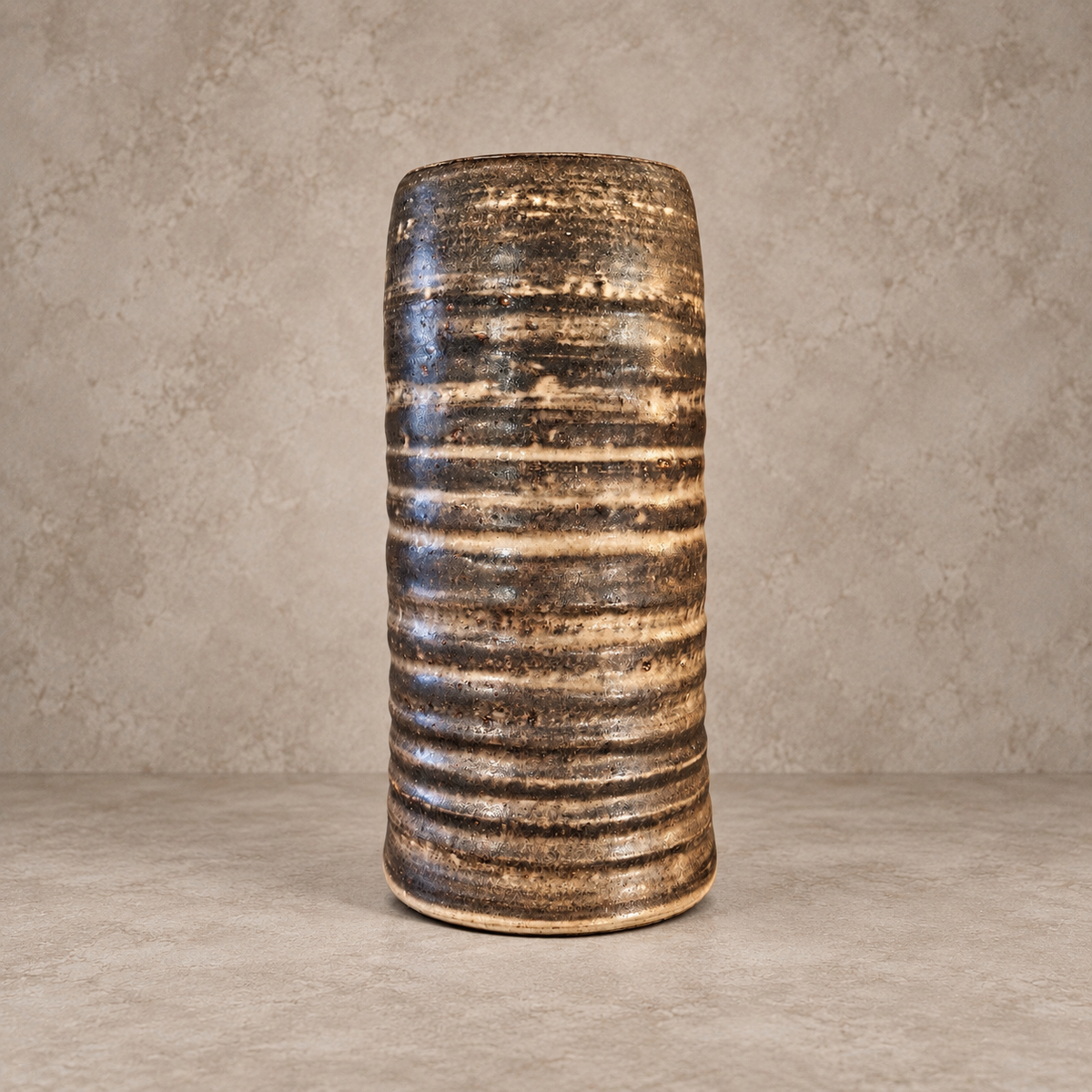 A tall, ribbed ceramic vase with a textured, horizontal striped pattern in earthy brown, beige, and black glazes, set against a neutral concrete-textured background.
