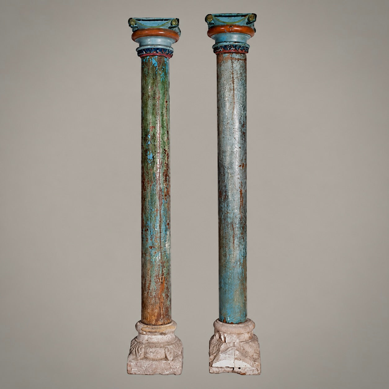 A pair of distressed, vintage-style classical column pedestals with peeling teal, green, and brown paint, set against a solid neutral gray background.