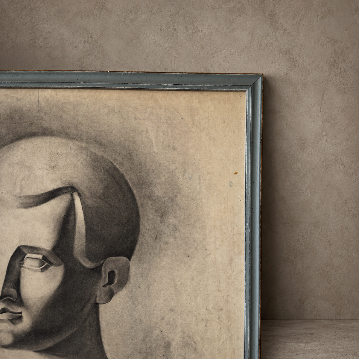 A framed monochrome portrait artwork leaning against a textured beige wall. The piece shows a stylized grayscale head in profile within a distressed blue-gray frame, with warm beige paper tones and dark charcoal shading.