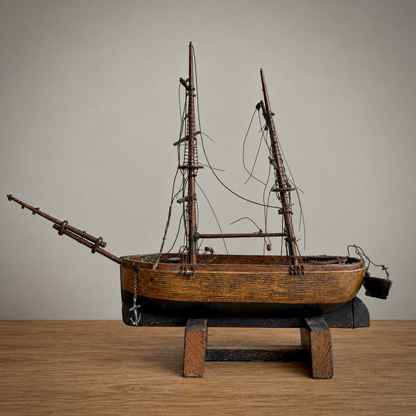 A detailed wooden model of a historic sailing ship with multiple masts, rigging, and an anchor, displayed on a wooden stand against a neutral background.