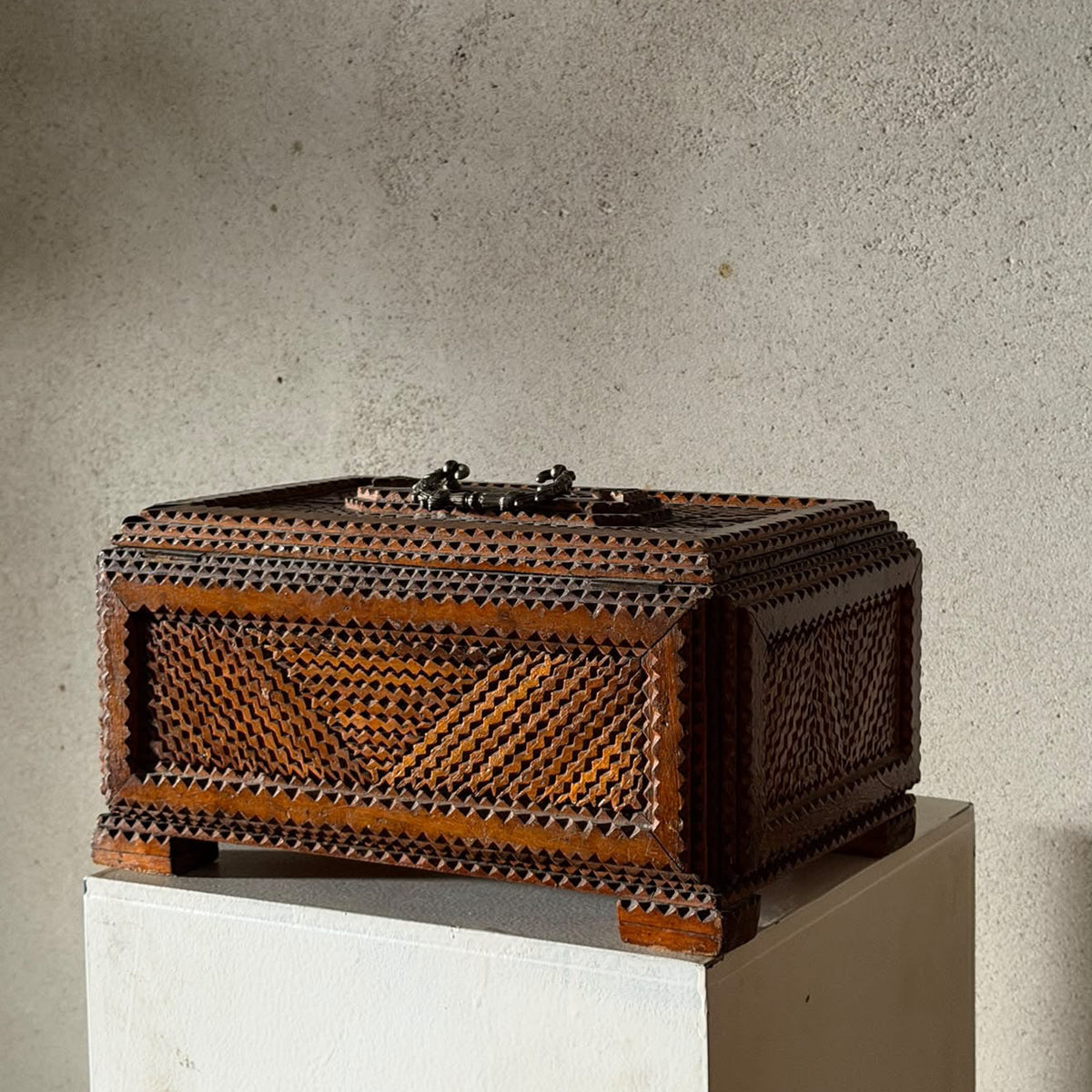 A dark brown carved wooden chest with intricate geometric lattice patterns, displayed on a white pedestal against a textured concrete wall.