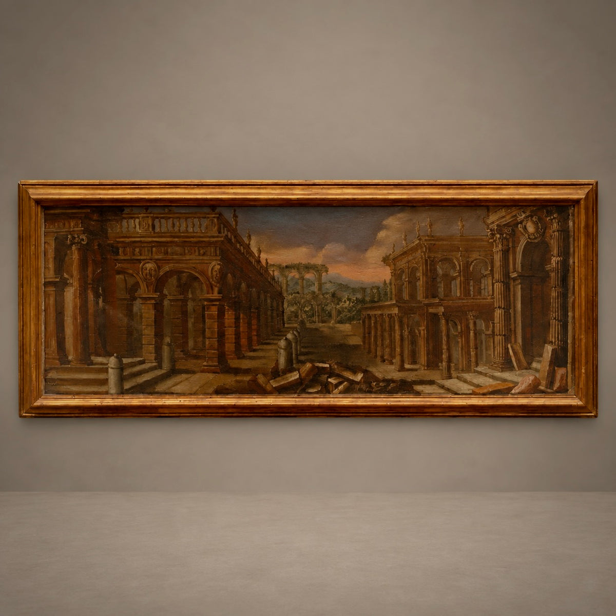 A framed classical landscape painting depicting ancient ruins with columns and arches under a dramatic sky, housed in a wide ornate gold frame and displayed against a neutral gray wall.