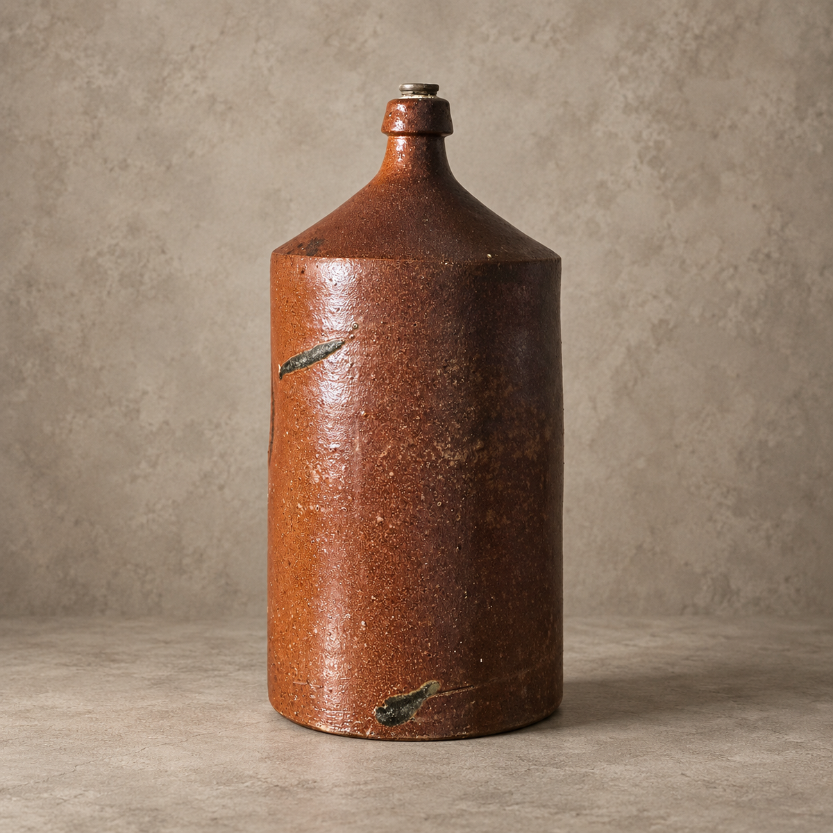 A rustic handmade stoneware bottle with a brown and terracotta glaze, featuring natural dark green crystalline inclusions, set against a textured neutral background.