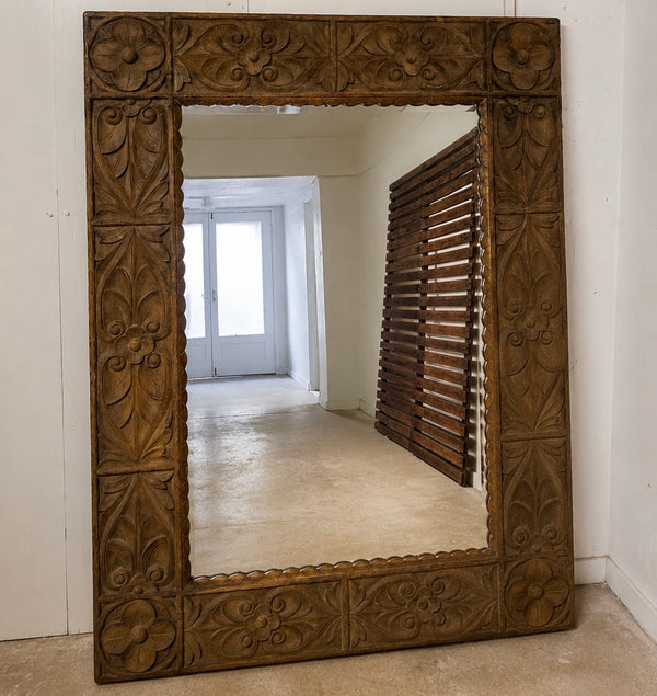 A large rectangular wall mirror with an ornate carved wooden frame, leaning against a white wall in a room. The mirror reflects a hallway with a slatted wooden door.