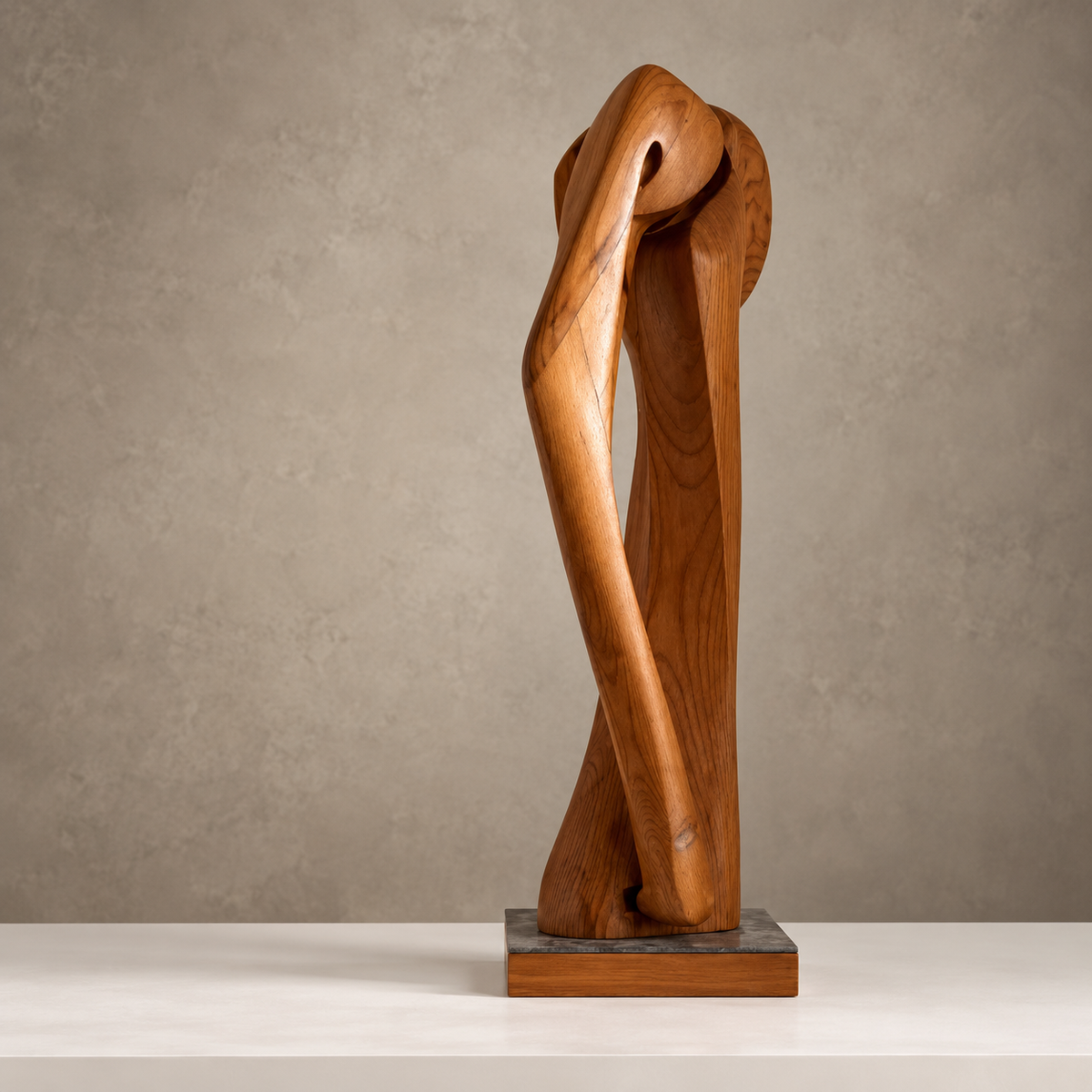 An abstract wooden sculpture with a polished medium-brown finish, featuring organic flowing lines and a rectangular dark stone base, displayed on a light surface against a textured gray wall.