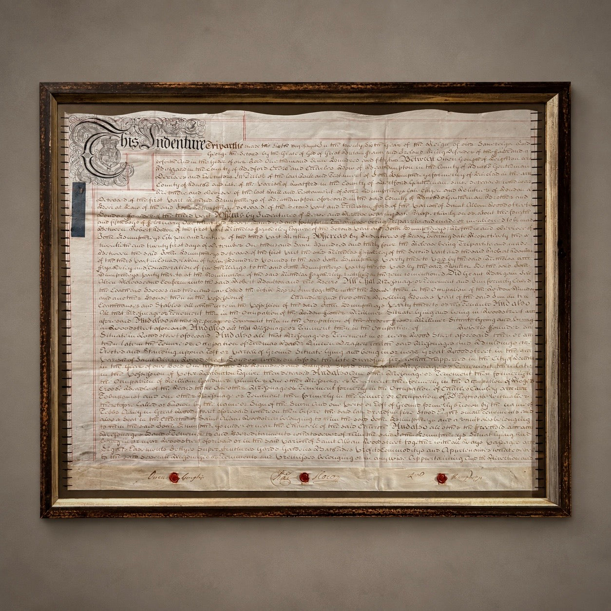 A framed property deed, featuring the original parchment document inside a large, ornate dark brown wooden frame., mounted in a dark wood frame against a neutral gray wall.