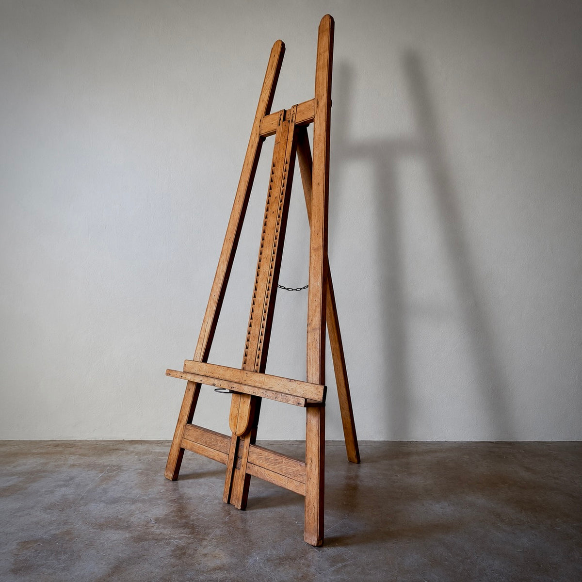 A vintage wooden A-frame easel with a height-adjustable central rack and a chain guard, standing on a concrete floor against a neutral gray wall.