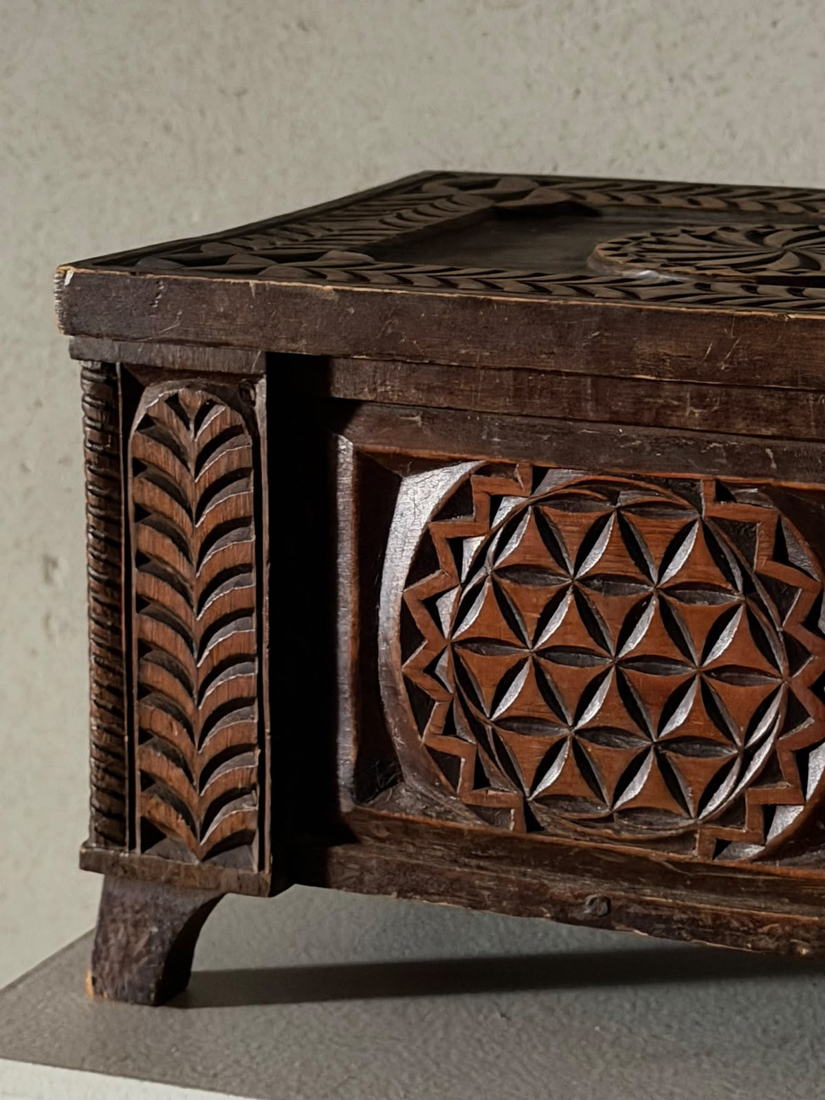 Close-up of a dark carved wooden chest or box with geometric and leaf-like relief patterns, shown against a neutral textured wall.