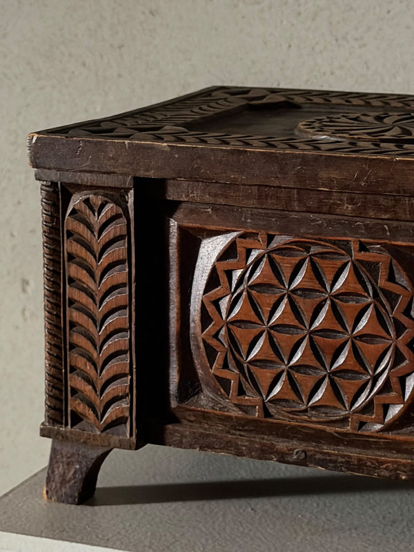 Close-up of a dark carved wooden chest or box with geometric and leaf-like relief patterns, shown against a neutral textured wall.