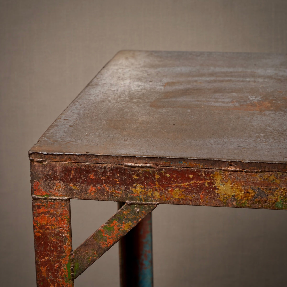 Close-up of a rustic industrial metal table corner with a weathered brown top and heavily distressed rusted red, orange, and green painted legs against a neutral gray background.