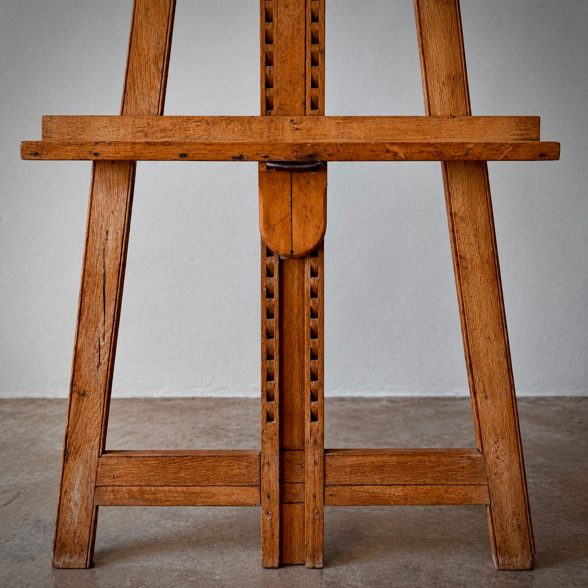 A close-up of a wooden artist easel, showing the center support and leg structure against a neutral gray wall and concrete floor.