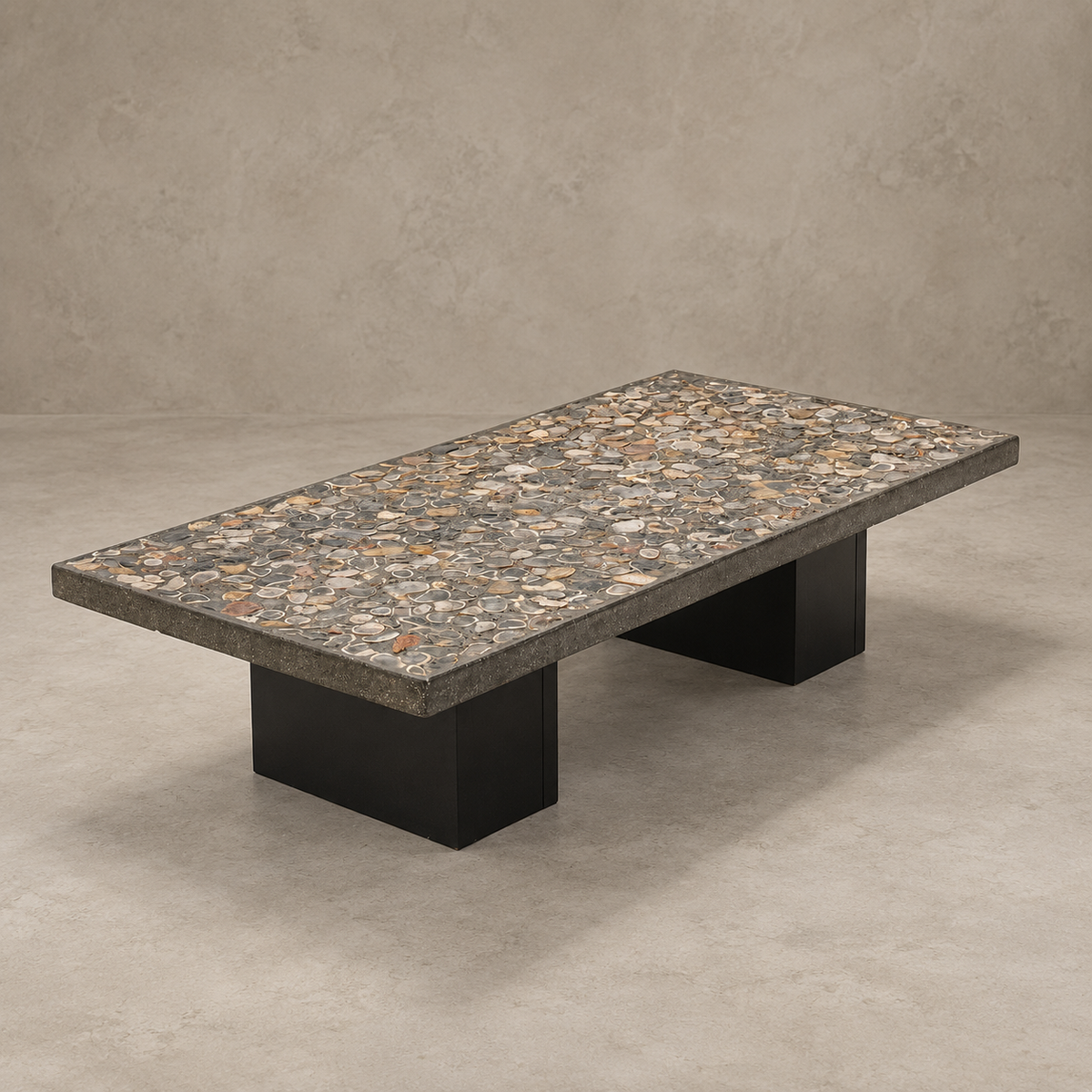 A rectangular coffee table with a speckled granite-look top in gray, beige, and brown tones, supported by two thick black block bases, set against a textured concrete wall and floor.