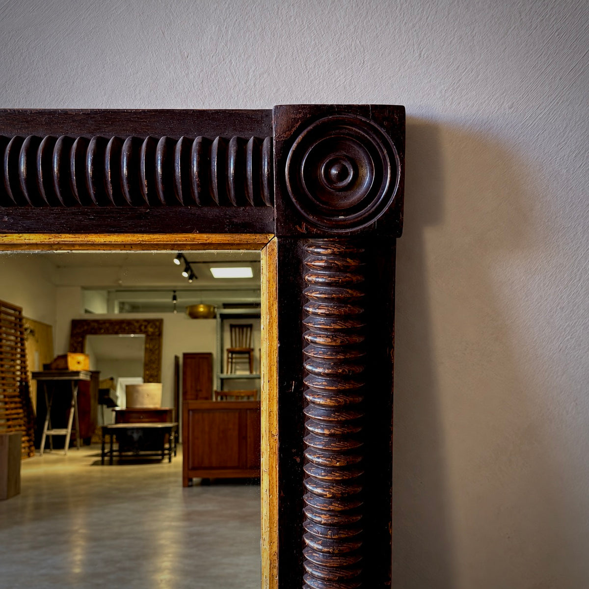 A close-up of a large antique-style mirror with an ornate dark brown carved wooden frame, set against a light gray wall with a reflection of an interior room visible in the mirror.