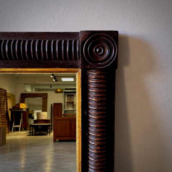 A close-up of a large antique-style mirror with an ornate dark brown carved wooden frame, set against a light gray wall with a reflection of an interior room visible in the mirror.