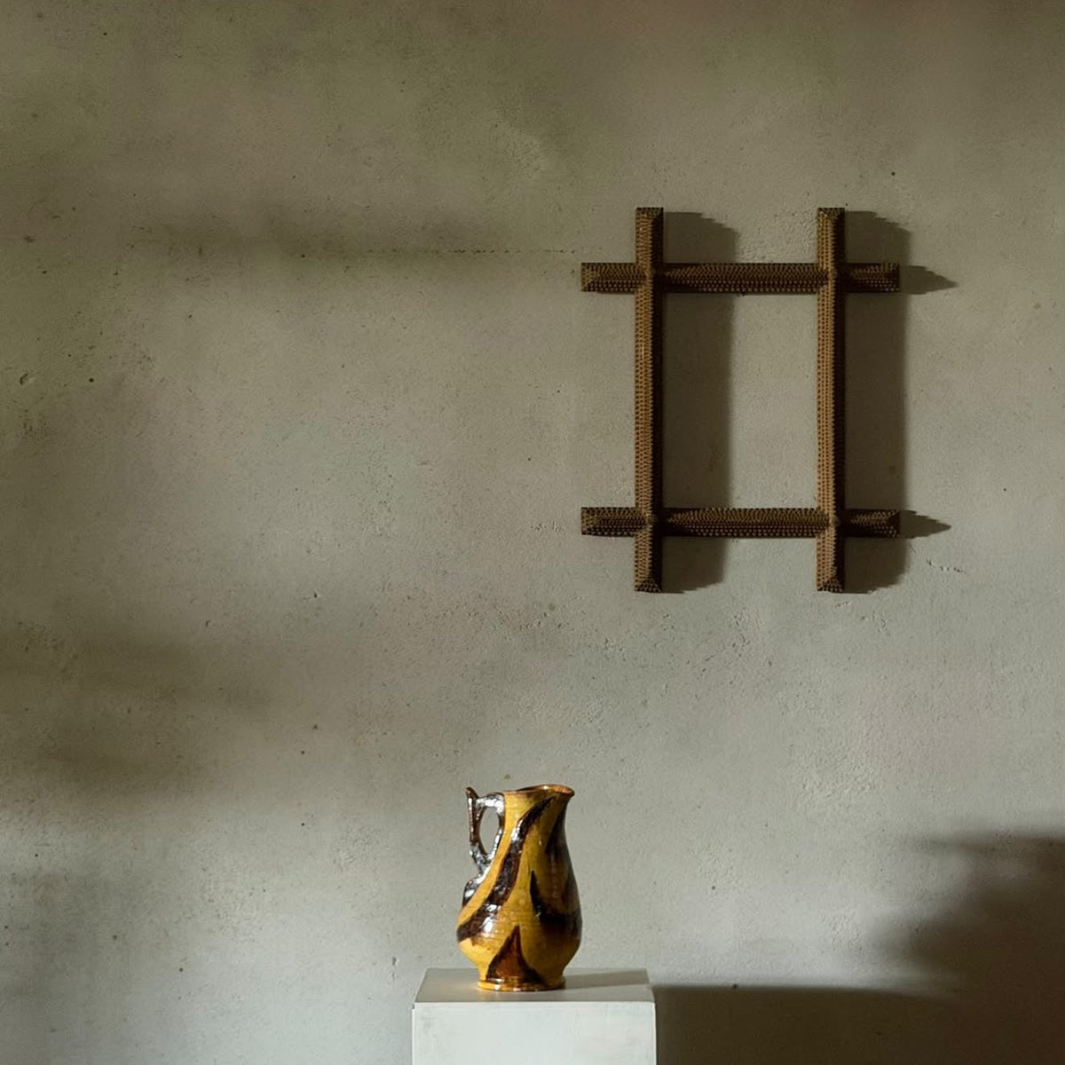A small yellow and black patterned ceramic pitcher sits on a white pedestal against a textured plaster wall, with a dark wooden wall-mounted frame above it.