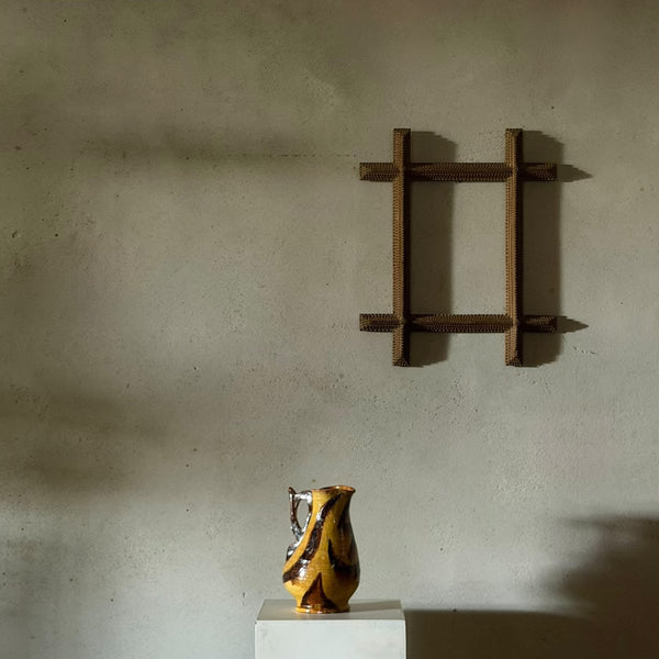 A small yellow and black patterned ceramic pitcher sits on a white pedestal against a textured plaster wall, with a dark wooden wall-mounted frame above it.