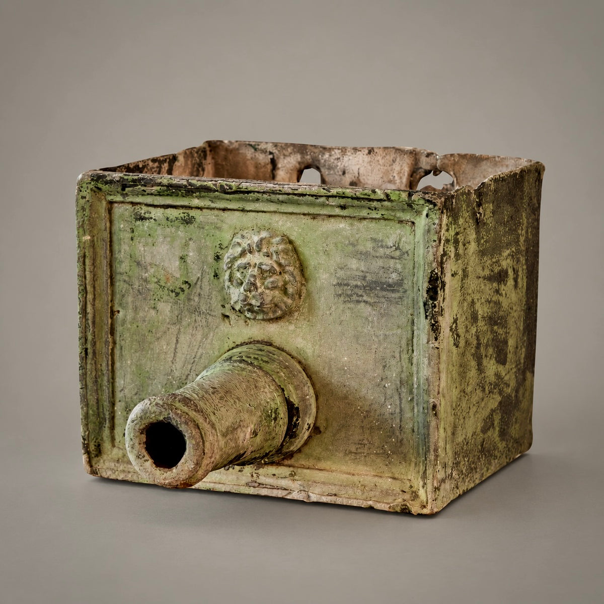 An ancient rectangular metal fountain head with a protruding cylindrical opening and a sculpted lion head motif on the front, shown against a neutral gray background.