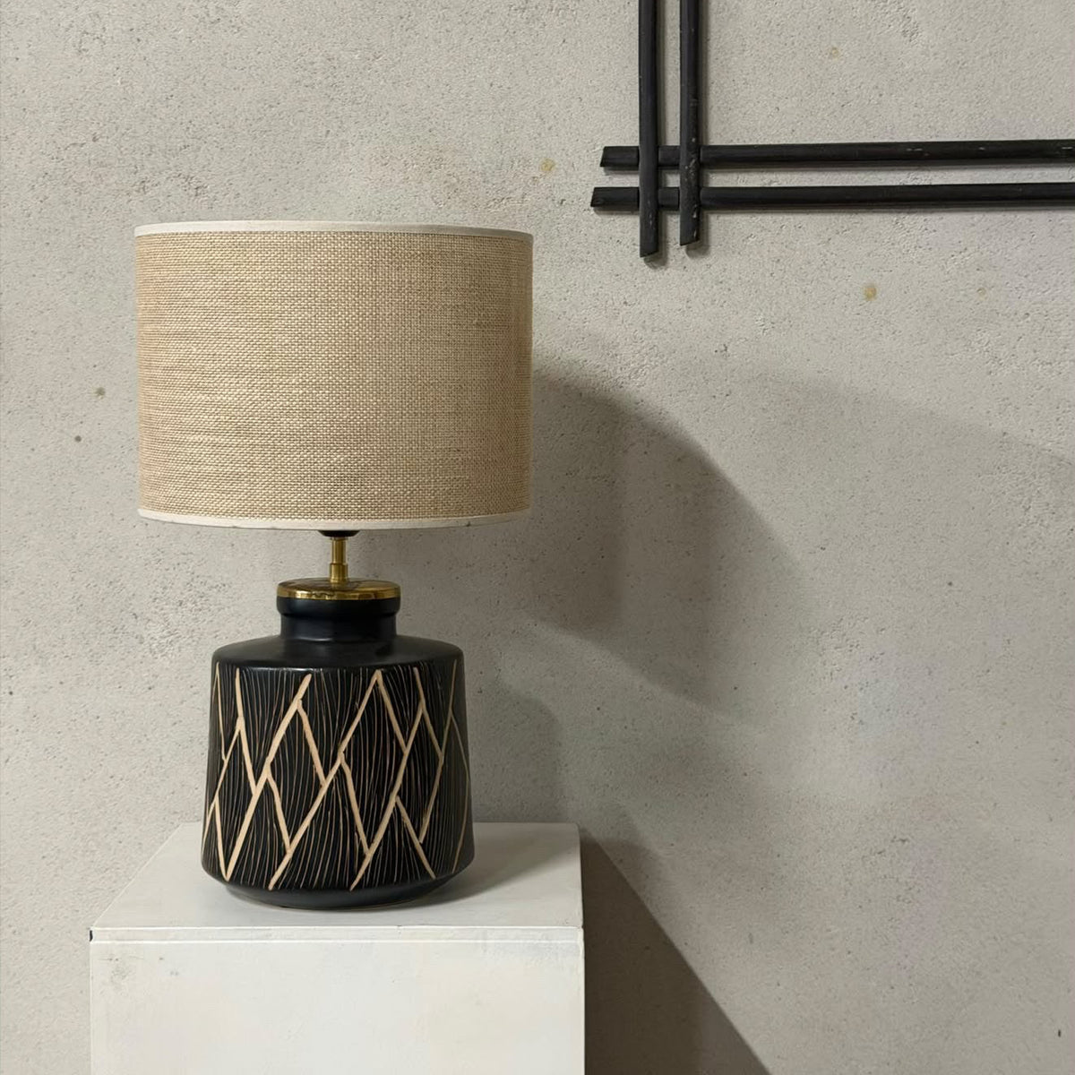 A table lamp with a beige linen drum shade and a black ceramic base featuring a gold geometric diamond pattern, displayed on a white pedestal against a textured gray wall.